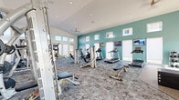 Located at 7755 Tierra del Sol Pkwy in Sparks, Nevada, this single-story fitness center is designed to support a comprehensive workout experience. The facility is divided into two distinct areas: a generously sized main gym floor and a bright, airy cycling studio.

The main gym area features a distinctive teal accent wall punctuated by a row of clerestory windows that flood the space with natural light. The patterned carpet flooring adds a modern aesthetic while providing comfort underfoot. This area is outfitted with a wide array of strength and cardio equipment, including treadmills, ellipticals, cable machines, selectorized weight machines, a multi-station functional trainer, free weights, and a plyo box for varied training routines. Mounted flat-screen televisions are positioned along the walls for entertainment during workouts.

The cycling studio offers a strikingly different atmosphere, featuring soaring vaulted ceilings, light hardwood flooring, and dramatic floor-to-ceiling windows that frame panoramic outdoor views of the surrounding Sparks landscape and distant hills. Multiple cycling bikes are arranged in the open space, which is bathed in natural light and serviced by a ceiling fan overhead. The minimalist, white-walled design of this room creates a clean, energizing environment ideal for focused training sessions.