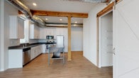 This single-floor loft-style unit offers a compelling mix of industrial character and modern design. The kitchen serves as a standout feature, showcasing white shaker cabinetry, dark countertops, and a full suite of stainless steel appliances including a dishwasher, range, microwave, and refrigerator. Exposed wooden beams and visible ductwork run overhead, adding a distinctive industrial aesthetic that carries throughout the open-plan living and kitchen area. Light hardwood-style flooring anchors the space and contributes to its bright, airy feel. A sliding barn door provides stylish separation between living areas, while large floor-to-ceiling windows and a glass sliding door open onto a private balcony, filling the space with natural light and offering outdoor views. The carpeted bedrooms are generously sized, with at least one bedroom featuring a window overlooking what appears to be a waterway. The primary bathroom includes a walk-in shower, wood-look tile flooring, and access to a walk-in closet with built-in shelving. A second full bathroom offers a soaking tub and shower combination. A dedicated laundry room with side-by-side washer and dryer units and overhead shelving adds everyday convenience. Throughout, the space balances warm, rustic elements with clean, contemporary finishes.