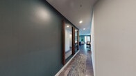 This thoughtfully designed single-floor residence combines modern aesthetics with functional living. A welcoming hallway, accented by rich walnut-framed mirrors and wood-look flooring, leads into the heart of the home. The open-concept living area features a striking teal accent wall, ceiling fan, large windows with patterned drapes, and ample natural light with outdoor views of surrounding greenery. Adjacent to the living space, the dining area benefits from expansive windows and sliding door access to an outdoor balcony, creating a bright and airy atmosphere. The kitchen is well-appointed with white flat-panel cabinetry, stainless steel appliances, a teal tile backsplash, quartz countertops, a center island with seating, and recessed lighting throughout. The primary bedroom showcases the same teal accent wall with large windows, plush carpeting, a ceiling fan, and an en-suite bathroom featuring a glass-enclosed shower, dual-sink vanity with dark countertops, and wood-look flooring. A second bedroom mirrors the cohesive design language with natural light and outdoor views. The second bathroom offers a full tub-shower combination, white cabinetry, and dark countertops. Throughout, the consistent use of teal accents, warm wood tones, and modern fixtures creates a unified and contemporary ambiance.