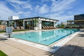 Located at 2055 Main St in Irvine, California, this expansive rooftop amenity level delivers a resort-style experience above the Irvine business district. The centerpiece is a generously sized lap pool with aqua-tiled lanes, flanked by lounge chairs and a large outdoor video screen — ideal for both recreation and entertaining. Adjacent to the pool, a spa with surrounding palm trees and tropical plantings creates a serene retreat atmosphere.

An outdoor kitchen and bar area feature granite-topped counters, built-in appliances, and a flat-top grill, seamlessly connected to the interior clubroom through large sliding glass doors. The indoor lounge offers tile flooring, warm gold-toned ceilings with decorative lighting, a full bar island with woven barstools, and multiple flat-screen TVs, all overlooking the pool deck through floor-to-ceiling glass.

The terrace extends into multiple distinct zones, including a sun-soaking lawn area with chaise lounges, a fire pit lounge with plush seating, and a landscaped garden promenade with ornamental grasses, succulents, and raised planters. String lights and open-sky views of the surrounding Irvine skyline complete the atmosphere, making this a well-appointed, multi-functional outdoor destination.