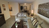 This single-floor residence offers a comfortable and cohesive living environment anchored by an open-concept layout that connects the living and dining areas seamlessly. The living room is furnished with a generously sized sectional sofa and a matching loveseat, both upholstered in soft gray fabric and accented with decorative pillows in muted earth tones. A dark-stained coffee table serves as a central focal point, complemented by a patterned area rug that adds warmth and texture to the carpeted floor. Ambient lighting is provided by a flush-mount ceiling fixture and a table lamp, creating a cozy and inviting atmosphere. The dining area, visible from the living space, features a dark wood dining table and chairs, accompanied by a decorative wall piece and a lush indoor plant that introduces a natural element to the interior. The neutral beige walls and light-toned carpet throughout the space contribute to a clean, understated aesthetic that feels both functional and welcoming. Wall art is thoughtfully placed to add visual interest without overwhelming the space. Overall, the layout promotes ease of movement and a relaxed, livable ambiance suited for everyday comfort.