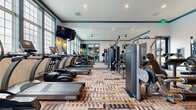 Located at 5056 Westerville Rd in Columbus, Ohio, this fitness center offers a thoughtfully designed workout space across a single floor. The room is outfitted with an extensive selection of TRUE brand equipment, including treadmills, stationary bikes, cable machines, and a variety of strength-training stations, providing a comprehensive range of exercise options. Large, multi-pane windows line the walls, allowing abundant natural light to fill the space and creating an open, airy atmosphere. Full-length mirrors further enhance the sense of depth and allow users to monitor their form during workouts. The ceiling features recessed lighting and a ceiling fan, ensuring consistent illumination and airflow throughout. A wall-mounted flat-screen TV provides entertainment during cardio sessions. The floor is covered in a vibrant multicolored patterned carpet that adds visual interest and a boutique-style aesthetic. The walls display textured wallcovering with a subtle geometric pattern, accented by a teal crown molding detail that runs along the perimeter. Sconce lighting and framed artwork contribute to the polished, upscale ambiance. A dedicated sanitation station with sanitizing wipes and supplies is conveniently positioned within the space. Glass doors connect the fitness center to an adjacent common area, enhancing accessibility and flow.