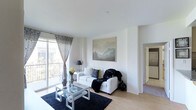 This single-floor residence offers a clean, contemporary design highlighted by an abundance of natural light and a neutral color palette throughout. The living area serves as the heart of the space, featuring smooth light hardwood flooring, crisp white walls, and high ceilings that contribute to an airy, open ambiance. A large sliding glass door flanked by elegant patterned curtains opens onto a private balcony, allowing natural light to fill the room and offering a pleasant outdoor connection. The living space is thoughtfully appointed, centered around a glass-top coffee table with a metallic frame set atop a deep navy area rug. A white sofa adorned with decorative pillows and a cozy throw anchors the seating arrangement, while a framed landscape painting depicting misty mountains adds a serene focal point to the wall. A doorway visible from the living area leads to an adjacent room, hinting at a well-connected and functional floor plan. A glimpse beyond reveals additional living spaces, including what appears to be a utility or laundry area. The overall atmosphere of the space is calm, modern, and welcoming, making it a comfortable and visually cohesive environment.