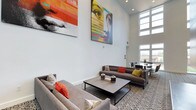 This single-floor space makes a striking first impression with its dramatic double-height ceilings, which amplify the sense of openness and grandeur throughout the main lounge area. The towering white walls serve as a gallery backdrop for two large-scale, vibrant artworks — bold, colorful portraits that command attention and establish a contemporary, artistic tone for the entire space.

The lounge area is thoughtfully arranged with modern gray sectional sofas accented by vivid orange, red, and yellow throw pillows, adding warmth and energy to the neutral palette. A low-profile rectangular coffee table anchors the seating arrangement, resting atop a bold leopard-print area rug that introduces a playful design element underfoot.

Natural light floods the space through tall windows positioned high along the walls and at the far end of the room, creating a bright, airy atmosphere. In the background, a dining or meeting area is visible, furnished with dark round tables and chairs, offering a functional secondary zone within the open-plan layout.

The overall ambiance is modern and curated, balancing artistic expression with comfortable, stylish furnishings in a spacious, well-lit environment.