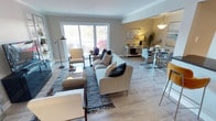 This single-floor space showcases an open-concept layout that integrates the living and dining areas into one cohesive, well-lit environment. Crown moulding along the ceiling adds a refined architectural touch, while light grey walls create a neutral, calming backdrop throughout. The living area is anchored by a generous light-toned sofa adorned with patterned throw pillows, complemented by an accent armchair and a textured area rug that defines the seating zone. A glass-top coffee table and woven ottoman add layers of texture and visual interest. Large sliding glass doors flood the space with abundant natural light and frame views of the surrounding outdoor landscape, enhancing the open and airy feel. Light wood-tone flooring runs continuously through the space, unifying the living and dining zones. The dining area features a round glass table with sleek modern chairs and is accented by a pendant light fixture overhead. A white sideboard along the wall provides functional storage while displaying decorative accessories, reinforcing the space's clean, contemporary aesthetic. Overall, the space balances comfort with modern sophistication.