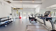 Located at 703 Whitney Avenue in New Haven, CT, this dedicated fitness center offers a clean, modern workout environment designed to support a full range of exercise routines. The single-floor space is defined by its bright, open layout, with white-paneled vaulted ceilings, exposed structural beams, and white support columns that lend an airy, spacious feel throughout.

Natural light pours in through multiple windows and glass-paneled doors along the perimeter, creating an inviting and energizing atmosphere. Large wall-mounted mirrors amplify the sense of space while allowing users to monitor their form during workouts. Track lighting overhead ensures the space remains well-lit at all hours.

The gym is comprehensively equipped for both cardio and strength training. The cardio zone features treadmills by TRUE Fitness, stationary bikes including a Peloton, and an elliptical machine. A dedicated strength area includes a multi-functional cable machine, an adjustable weight bench, and a full rack of dumbbells. A TRX suspension training rig is mounted to the ceiling beams, while a medicine ball rack adds options for functional fitness. The durable rubber flooring throughout provides comfort and safety underfoot. A staircase near the entrance suggests access to an upper level or adjacent area, rounding out this thoughtfully designed fitness facility.