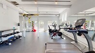 Located at 703 Whitney Avenue in New Haven, CT, this dedicated fitness center offers a clean, modern workout environment designed to support a full range of exercise routines. The single-floor space is defined by its bright, open layout, with white-paneled vaulted ceilings, exposed structural beams, and white support columns that lend an airy, spacious feel throughout.

Natural light pours in through multiple windows and glass-paneled doors along the perimeter, creating an inviting and energizing atmosphere. Large wall-mounted mirrors amplify the sense of space while allowing users to monitor their form during workouts. Track lighting overhead ensures the space remains well-lit at all hours.

The gym is comprehensively equipped for both cardio and strength training. The cardio zone features treadmills by TRUE Fitness, stationary bikes including a Peloton, and an elliptical machine. A dedicated strength area includes a multi-functional cable machine, an adjustable weight bench, and a full rack of dumbbells. A TRX suspension training rig is mounted to the ceiling beams, while a medicine ball rack adds options for functional fitness. The durable rubber flooring throughout provides comfort and safety underfoot. A staircase near the entrance suggests access to an upper level or adjacent area, rounding out this thoughtfully designed fitness facility.