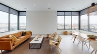 This single-floor space showcases a thoughtfully designed open-plan layout that seamlessly integrates a living area and a contemporary kitchen. The living area is defined by large, floor-to-ceiling windows with black frames on two walls, flooding the space with natural light while offering expansive views of the surrounding urban skyline. Light-toned large-format floor tiles run throughout, contributing to a clean, airy atmosphere. The seating arrangement features a tan leather sofa paired with natural rattan chairs and a minimalist marble-top coffee table, all anchored by a woven area rug.

The kitchen is finished to a high standard, featuring flat-front cabinetry in a warm, light wood tone paired with light stone countertops. A striking mosaic tile backsplash adds visual texture behind the sink and appliances, which include stainless steel finishes throughout. A substantial natural wood island anchors the kitchen, illuminated by matte black pendant lights overhead, complementing the modern yet warm aesthetic of the space.

Overall, the space presents a cohesive, minimalist design language with warm natural material accents, generous natural light, and a well-considered flow between functional zones, creating a bright and sophisticated environment.
