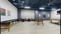 This single-floor game and lounge space offers a generously sized, open-plan layout designed for recreation and social gathering. The room is anchored by an exposed black ceiling with industrial ductwork and overhead linear lighting, lending an urban, loft-like aesthetic. Light wood-plank flooring runs throughout, providing a warm contrast to the dark charcoal accent walls.

A striking checkered feature wall serves as the focal point of the lounge seating area, where a long gray sectional sofa is paired with bold rust-orange accent chairs and small side tables — ideal for relaxed conversation. Adjacent to this seating zone, a high-top table with bar stools provides an additional casual gathering spot.

The space is well-equipped for entertainment, featuring foosball tables, a shuffleboard table, and a pool table placed across the open floor. A separate seating nook along one wall showcases a series of angled mirror panels, comfortable lounge chairs, and potted greenery, creating a more intimate retreat within the larger room. A wall-mounted flat-screen television is installed above an artistic mural of abstract circular line work on a white paneled wall. Colorful framed artwork adds personality to another corner. The overall ambiance is modern, energetic, and thoughtfully designed for both active play and casual lounging.