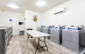 Located at 255 E Grand Ave in Rahway, New Jersey, this shared laundry facility is a thoughtfully designed, single-floor space built to accommodate residents with efficiency and comfort in mind. The room features light-toned tile flooring and bright white walls, creating a clean and welcoming environment illuminated by flush-mount ceiling lights and a wall-mounted mini-split air conditioning unit for year-round comfort.

The facility is equipped with a row of top-loading washing machines along one wall and a separate row of front-loading washers along another, providing ample capacity for multiple users. A large folding table positioned centrally in the room offers generous space for sorting and folding laundry. A decorative wrought iron bench provides seating while waiting, accompanied by a multi-tiered wooden shelf holding plants, books, and small decorative items that add a homey touch to the space.

The walls are accented with framed artwork, a decorative laundry-themed sign, a wall clock, and green wreaths, contributing to a warm and organized ambiance. A utility sink is also present, adding functional convenience. The red entry door provides a clear focal point and easy access. Overall, the space balances practical utility with a clean, inviting aesthetic suitable for everyday residential use.