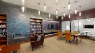 Located in Philadelphia's vibrant Fairmount neighborhood, this thoughtfully designed amenity space offers a seamless blend of functionality and contemporary style. The layout is anchored by a spacious conference/dining area featuring a long wooden table surrounded by warm amber upholstered chairs, lit by a row of sleek cylindrical pendant lights that hang gracefully overhead. Glass-walled private meeting rooms with flat-screen displays and colorful seating are positioned adjacent, providing versatile options for focused work or collaboration. A defining architectural feature throughout is the series of vertical wood slat partitions that elegantly divide zones while allowing natural light and outdoor views to filter through floor-to-ceiling glazing, connecting the interior to a lush exterior terrace. The lounge area offers a welcoming retreat with rich brown leather armchairs, a stone-tiled fireplace flanked by built-in wooden bookshelves stocked with curated reading material, and a flat-screen display set within a deep-hued accent wall. Warm hardwood flooring, muted taupe and plum wall tones, and carefully placed pendant lighting contribute to an inviting, upscale ambiance that balances professional utility with relaxed comfort throughout the space.