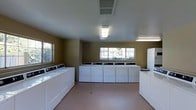 This single-floor shared laundry facility is designed to accommodate high-capacity use with efficiency and practicality in mind. The room is outfitted with a generous number of commercial-grade top-loading washing machines and front-loading dryers arranged along the walls, providing ample capacity for multiple users simultaneously. The warm tan-painted walls give the space a neutral, clean aesthetic, while the smooth, easy-to-maintain flooring in a soft terracotta tone runs throughout the room. Overhead fluorescent lighting ensures the space is well-lit at all times, complemented by natural light streaming in through multiple windows positioned on two walls. The windows also offer views to the surrounding outdoor environment, contributing to an open and airy feel. A centrally located HVAC vent in the ceiling supports comfortable air circulation year-round. The layout is thoughtfully organized to allow users to move freely between machines, with sufficient open floor space for sorting and managing laundry. Overall, this laundry facility is a functional, no-frills communal space built to serve the practical needs of its users effectively.