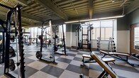 Located at 707 S Fourth St in Champaign, IL, this fitness center offers a comprehensive, single-floor workout facility designed to accommodate a wide range of exercise needs. The space features a bold checkered tile floor in alternating shades of gray, providing a striking visual contrast throughout the main exercise area.

The strength training section is extensively equipped with cable machines, squat racks, Smith machines, free weight stations, and a full rack of dumbbells, all reflected in large wall-mounted mirrors that enhance the sense of depth and space. The cardio zone houses a lineup of Life Fitness ellipticals, stair climbers, and treadmills, positioned near expansive floor-to-ceiling windows that allow abundant natural light and offer views of the surrounding Champaign streetscape.

The exposed concrete ceiling with visible structural beams and ductwork gives the facility an industrial character, complemented by sage green accent walls and efficient linear lighting. A dedicated hardwood floor area transitions seamlessly from the main workout space, and open shelving along one wall provides convenient storage. A dramatic open staircase with sleek metal railings and glass panels, flanked by a floor-to-ceiling window wall, serves as a striking architectural focal point connecting the fitness floor to adjacent levels of the building.