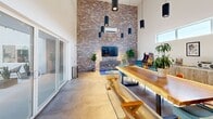 This single-floor space makes a striking impression through its dramatic architectural contrasts and thoughtful design. The defining feature of the main area is a soaring ceiling height that amplifies the sense of space well beyond its modest footprint. A full-height exposed brick accent wall anchors the room, paired with crisp white walls and polished concrete flooring to create an industrial-modern aesthetic. A row of clerestory windows lines the upper wall, drawing in natural light and enhancing the open, airy feel. Cylindrical black pendant lights hang at varying heights, adding focused illumination and a contemporary edge.

The open layout accommodates a live-edge wood communal table with bench seating, a lounge area, and additional accent chairs, making the space versatile for both collaborative and social use. Large sliding glass doors on one side offer connectivity to an adjoining area.

The kitchen runs along one wall and features rustic wood-grain cabinetry, a white quartz countertop, white subway tile backsplash, a gold double undermount sink, a stainless steel refrigerator, and a floating display shelf—blending functionality with refined detailing throughout.
