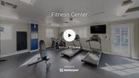 Located at 1025 South Gilbert Street in Hemet, California, this fitness center is a dedicated single-room exercise facility designed to support a well-rounded workout routine. The space is finished with a durable dark rubber floor, smooth white ceilings with crown molding, and neutral gray walls accented by bold blue horizontal stripe detailing, lending the room a clean, energetic aesthetic.

Natural light enters through multiple windows along the perimeter, supplemented by overhead fluorescent fixtures that ensure the space remains bright throughout the day. A large wall-mounted flat-screen television provides entertainment during workouts, while full-length mirrors along one wall visually expand the room and allow for form monitoring.

The cardio area is equipped with multiple treadmills and an elliptical cross-trainer, arranged to maximize floor space. A multi-functional cable machine anchors the strength-training section, providing options for a wide variety of resistance exercises. Additional equipment includes a weight bench, free weights, and a wall-mounted padded mat for stretching or bodyweight exercises. A posted rules and information board near the entrance keeps users informed of facility guidelines. French doors offer convenient access and contribute to the room's openness. Overall, the facility is thoughtfully equipped to serve the fitness needs of its users in a compact, functional environment.