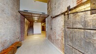 This distinctive single-floor space masterfully combines historic industrial character with contemporary functionality. Exposed brick walls throughout speak to the building's storied past, while polished and raw concrete floors ground the space with a robust, utilitarian aesthetic. Soaring ceiling heights in the main corridor create a dramatic sense of openness, enhanced by a mezzanine level overhead supported by a warm wood-paneled soffit with recessed track lighting. A set of stairs leads up to the mezzanine area, which offers an additional layer of usable space above.

A heavy-duty industrial sliding metal door — a nod to the building's original purpose — serves as a striking architectural feature at the entrance corridor, adding authenticity and visual interest. The common areas are clean and well-lit, featuring modern fluorescent strip lighting, neutral white walls, and polished terrazzo-style concrete floors that contrast beautifully with the raw brick. A stainless steel elevator provides convenient vertical access, accompanied by a bank of individual mailboxes along the hallway wall. Exposed ductwork on the ceiling adds to the industrial charm. A dedicated bike storage room lined with wall-mounted bike racks provides practical amenity. Together, these elements create a dynamic, loft-like environment rich in texture and character.