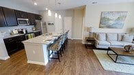 This single-floor residence showcases an open-concept layout that effortlessly connects the kitchen, dining, and living areas into one cohesive living space. The kitchen is equipped with stainless steel appliances, dark espresso cabinetry, and a generous granite-topped island that doubles as a breakfast bar, accommodating multiple bar-height chairs with patterned upholstery. Pendant lighting hangs above the island, adding a refined, contemporary touch to the space. The kitchen flows naturally into a casual dining area and an adjacent living room, creating an inviting environment ideal for both entertaining and everyday living. Throughout the main living area, rich hardwood-style flooring adds warmth and continuity, tying the spaces together visually. Neutral-toned walls provide a clean, versatile backdrop that complements the darker cabinetry and furnishings. The living area is comfortably appointed with a sofa, side table, and lamp, conveying a relaxed yet polished ambiance. A white interior door is visible toward the rear of the space, suggesting additional private rooms beyond the open living area.