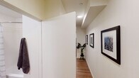 This well-appointed single-floor studio offers a thoughtfully designed layout that maximizes functionality and comfort. The main living area serves as the heart of the space, featuring warm hardwood-style flooring, neutral white walls, and recessed lighting that creates a bright, welcoming ambiance. A sofa, a compact dining table with chairs, and a round decorative mirror contribute to the room's modern aesthetic, while a window allows natural light to filter in. A large sliding-door closet provides ample storage directly within the living area.

The kitchen is efficiently designed with light wood cabinetry, white countertops, and a full suite of stainless steel appliances including an electric range, over-the-range microwave, dishwasher, and refrigerator — offering everything needed for everyday cooking.

A well-lit hallway, decorated with framed artwork and accented by a potted plant, connects the main living space to the rest of the unit. The hallway leads to a clean, modern bathroom featuring a white vanity with an undermount sink, a full tub with a shower curtain, a toilet, and warm wood-tone flooring that complements the rest of the unit. The overall design is cohesive and contemporary, with a neutral color palette and minimalist touches throughout.