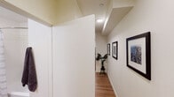 This well-appointed single-floor studio offers a thoughtfully designed layout that maximizes functionality and comfort. The main living area serves as the heart of the space, featuring warm hardwood-style flooring, neutral white walls, and recessed lighting that creates a bright, welcoming ambiance. A sofa, a compact dining table with chairs, and a round decorative mirror contribute to the room's modern aesthetic, while a window allows natural light to filter in. A large sliding-door closet provides ample storage directly within the living area.

The kitchen is efficiently designed with light wood cabinetry, white countertops, and a full suite of stainless steel appliances including an electric range, over-the-range microwave, dishwasher, and refrigerator — offering everything needed for everyday cooking.

A well-lit hallway, decorated with framed artwork and accented by a potted plant, connects the main living space to the rest of the unit. The hallway leads to a clean, modern bathroom featuring a white vanity with an undermount sink, a full tub with a shower curtain, a toilet, and warm wood-tone flooring that complements the rest of the unit. The overall design is cohesive and contemporary, with a neutral color palette and minimalist touches throughout.