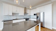 Located at 1414 Broadview Ave in Columbus, Ohio, this single-story home blends modern design with functional living. The kitchen serves as a standout centerpiece, showcasing crisp white shaker-style cabinetry that extends to the ceiling and is accented by elegant crown molding. A generously sized central island with a sleek dark quartz countertop provides ample workspace and casual seating, while matching counters line the perimeter of the room. A marble-toned subway tile backsplash adds texture and sophistication, complementing the overall clean aesthetic. Stainless steel appliances, including a French door refrigerator with an ice and water dispenser, a built-in microwave, and a professional-grade range with a wall-mounted stainless hood, deliver a fully equipped cooking environment. Two clear glass pendant lights hang above the island, casting a warm, inviting glow that pairs well with the recessed lighting overhead. Warm hardwood flooring grounds the space and flows through the home, adding natural warmth to the contemporary design. The thoughtful layout and high-quality finishes throughout make this home a well-appointed and visually cohesive residence in the heart of Columbus.