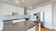 Located at 1414 Broadview Ave in Columbus, Ohio, this single-story home blends modern design with functional living. The kitchen serves as a standout centerpiece, showcasing crisp white shaker-style cabinetry that extends to the ceiling and is accented by elegant crown molding. A generously sized central island with a sleek dark quartz countertop provides ample workspace and casual seating, while matching counters line the perimeter of the room. A marble-toned subway tile backsplash adds texture and sophistication, complementing the overall clean aesthetic. Stainless steel appliances, including a French door refrigerator with an ice and water dispenser, a built-in microwave, and a professional-grade range with a wall-mounted stainless hood, deliver a fully equipped cooking environment. Two clear glass pendant lights hang above the island, casting a warm, inviting glow that pairs well with the recessed lighting overhead. Warm hardwood flooring grounds the space and flows through the home, adding natural warmth to the contemporary design. The thoughtful layout and high-quality finishes throughout make this home a well-appointed and visually cohesive residence in the heart of Columbus.