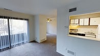 This single-floor space features an open-concept living and dining area finished with wall-to-wall carpeting and neutral-toned walls that create a clean, understated backdrop. Natural light enters through a large sliding glass door fitted with vertical blinds, brightening the main living area. A ceiling fan is centrally positioned overhead, providing both air circulation and a subtle decorative element. Floor vents are visible throughout, indicating a forced-air climate control system, and a wall-mounted thermostat adds to the functional character of the space. A defining architectural feature is the pass-through window connecting the living area to the kitchen, which enhances the sense of openness between the two spaces. The kitchen, visible through this opening, includes an overhead range hood with under-cabinet lighting, white upper cabinetry, and a gas or electric range, providing a compact yet well-equipped cooking area. The layout flows naturally from the front living zone through to a secondary living or dining area beyond a partial wall, allowing for versatile furniture arrangement. Overall, the space presents a practical and well-organized single-level layout suited for everyday living.