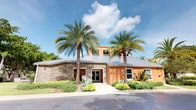 This single-story building presents a visually distinctive exterior that combines natural stone cladding with warm terracotta-hued stucco walls, creating an appealing contrast that is both inviting and architecturally refined. A standing-seam metal roof crowns the structure, adding a modern, durable finish that complements the overall design. The entrance is framed by glass doors and flanked by two towering palm trees, which lend a tropical, resort-like character to the property. Carefully maintained landscaping surrounds the building, featuring low-growing shrubs, ornamental grasses, and tropical plantings that enhance the curb appeal and reinforce the warm-climate setting. A raised central section above the main roofline adds visual interest and architectural depth to the facade, incorporating additional window openings for light. The clean, well-maintained walkway and driveway approach contribute to an organized and professional exterior presentation. The overall ambiance of the building is one of a well-appointed, tropical commercial or community-use space, designed to feel welcoming while maintaining a polished and functional appearance suited to its sunny, coastal surroundings.