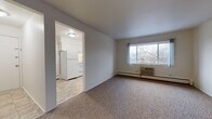 This two-bedroom, one-bathroom unit in Ann Arbor, Michigan offers a straightforward single-floor layout designed for practical, comfortable living. The living area is carpeted and receives natural light through a large picture window that frames views of surrounding trees. A wall-mounted air conditioning unit and baseboard heater provide year-round climate control. An open rectangular archway connects the living space directly to the kitchen, which features tile flooring, white cabinetry extending to the ceiling, a white refrigerator, electric range, and a double-basin sink positioned beneath a window. The layout provides ample counter and storage space for everyday use. Both bedrooms are generously proportioned, finished with neutral carpet, and equipped with baseboard heating. One bedroom benefits from two windows on adjoining walls, filling the room with natural light and outdoor views, while the other includes access to a dedicated closet space. The full bathroom is finished with tile flooring, white subway-style wall tile surrounding the tub and shower, a vanity with under-sink storage, a mirrored medicine cabinet, and a window for natural light and ventilation. Throughout the unit, the neutral color palette and consistent finishes create a clean, cohesive atmosphere.