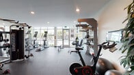 This fitness center offers a comprehensive workout environment designed with both functionality and comfort in mind. The space is defined by expansive floor-to-ceiling windows along one full wall, flooding the interior with natural light and providing unobstructed views of an outdoor pool and landscaped surroundings. Direct access to the pool area is available through glass-paneled doors, creating a seamless connection between indoor and outdoor amenities.

The flooring throughout consists of durable rubber gym matting, providing cushioning and traction suitable for a wide range of exercises. The cardio zone features multiple treadmills, elliptical trainers, and stair climbers, several equipped with personal display screens. A dedicated TRX Training Zone wall-mounted rack organizes suspension trainers, medicine balls, and kettlebells for functional fitness training.

Strength training is well-represented through a multi-station cable machine, adjustable weight benches, a dumbbell rack, and additional resistance equipment. A spin bike with a connected display screen adds cycling options. Recessed lighting in the smooth ceiling supplements abundant natural light. A separate interior alcove with louvered doors provides additional storage or utility access, completing this well-rounded facility.
