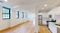 This thoughtfully designed single-floor space offers a clean, modern aesthetic highlighted by light hardwood flooring that runs seamlessly throughout every room. The open-concept layout connects the living area and kitchen, creating a sense of spaciousness and easy flow between functional zones. Crisp white walls and recessed lighting keep the interior bright and airy.

The kitchen is well-appointed with sleek, high-gloss white cabinetry, a full suite of stainless steel appliances including a refrigerator, gas range, built-in microwave, and dishwasher, along with ample counter and storage space. A structural column adds a subtle architectural character to the kitchen-living transition.

The living area is a standout, featuring an electric fireplace set into a gray accent niche with a warm wood mantle, flanked by black-framed grid windows that invite generous natural light and outdoor views. Additional rooms provide flexible space, each finished with the same consistent hardwood flooring and neutral white walls. The bathroom is clean and functional, featuring a full tub-shower combination, toilet, and vanity, all finished in white. Throughout, the black-framed windows create a striking contrast against the bright interiors, lending the space a contemporary character.