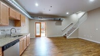 This two-story unit blends industrial character with practical residential design. The main level opens into a generous living and dining area finished with warm wood-look flooring throughout. Exposed cylindrical ductwork runs along the ceiling, adding an industrial aesthetic that defines the space's character. Recessed lighting illuminates the open floor plan, while a wood-framed glass door at the rear provides access to the outdoors and draws in natural light. A white-railed staircase leads to the upper level.

The kitchen is well-appointed along a single wall, featuring warm honey-toned cabinetry, granite countertops, a tile backsplash with decorative mosaic accents, and a full suite of black and stainless steel appliances including a refrigerator, range, built-in microwave, and dishwasher. A utility room on the main level houses a stacked washer and dryer alongside a water heater and wire shelving for additional storage.

Upstairs, carpeted bedrooms offer a quieter retreat, each with closet space and access to natural light. The upper level also features a full bathroom with a tub-shower combination and wood-look flooring, along with a vanity with warm wood cabinetry and a white countertop.
