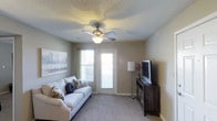 This single-floor space offers a welcoming and functional layout designed for everyday comfort. The living room serves as the heart of the space, featuring plush carpeted flooring and warm, neutral-toned walls that create a calm and inviting atmosphere. A ceiling fan with an integrated light fixture is centrally mounted on the textured ceiling, providing both air circulation and ambient lighting throughout the room. Natural light flows generously into the space through two windows fitted with blinds, as well as a glass-paneled door that leads to an exterior area, further brightening the interior. The living room is thoughtfully arranged to accommodate a sofa with decorative pillows, positioned to face an entertainment setup that includes a flat-screen television resting on a dark wood media console. A tall floor lamp beside the console adds supplementary lighting and a touch of elegance. A landscape wall art piece adorns the wall, contributing to the space's understated, homey aesthetic. The overall ambiance is clean, functional, and comfortable, making it well-suited for relaxing and everyday living.