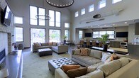 This two-story space makes an immediate impression with its soaring double-height ceilings, which are accentuated by an abundance of natural light streaming through dramatic floor-to-ceiling windows along the main wall and a second row of clerestory windows above. The lofty volume is anchored by a statement chandelier with an exposed bulb ring design, complemented by a smaller matching pendant, lending an industrial-modern aesthetic to the interior.

The main lounge area is generously proportioned, featuring multiple large sectional sofas in neutral gray tones, dressed with accent pillows in warm oranges and earth tones. A tufted ottoman serves as a central focal point, providing both style and function. The light-toned area rug grounds the seating arrangement and adds texture to the space.

Transitioning deeper into the layout, a defined dining and bar area is visible, complete with a long counter-height surface, upholstered bar stools, and a wall-mounted television. Large windows in this zone continue the theme of natural light. Potted tropical plants are distributed throughout, adding warmth and life. The overall ambiance is modern, open, and welcoming.