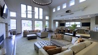 This two-story space makes an immediate impression with its soaring double-height ceilings, which are accentuated by an abundance of natural light streaming through dramatic floor-to-ceiling windows along the main wall and a second row of clerestory windows above. The lofty volume is anchored by a statement chandelier with an exposed bulb ring design, complemented by a smaller matching pendant, lending an industrial-modern aesthetic to the interior.

The main lounge area is generously proportioned, featuring multiple large sectional sofas in neutral gray tones, dressed with accent pillows in warm oranges and earth tones. A tufted ottoman serves as a central focal point, providing both style and function. The light-toned area rug grounds the seating arrangement and adds texture to the space.

Transitioning deeper into the layout, a defined dining and bar area is visible, complete with a long counter-height surface, upholstered bar stools, and a wall-mounted television. Large windows in this zone continue the theme of natural light. Potted tropical plants are distributed throughout, adding warmth and life. The overall ambiance is modern, open, and welcoming.