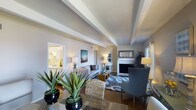 Located at 890 Quail Lake Circle in the desirable Quail Lake area of Colorado Springs, Colorado, this single-story residence offers a thoughtfully designed living environment with refined architectural details. The living room immediately captures attention with its vaulted ceiling accented by exposed beams, creating a sense of openness and character. Rich hardwood flooring extends throughout, adding warmth and continuity to the space. A classic fireplace with a decorative mantle serves as the focal point of the living area, flanked by comfortable seating and complemented by carefully chosen furnishings, including woven accent chairs and layered area rugs that add texture and depth. Natural light filters in through the windows, brightening the neutral, warm-toned walls and enhancing the inviting ambiance. Lush indoor plants add a vibrant, organic touch to the interior. A glimpse into the adjacent kitchen reveals bright cabinetry and a well-appointed layout. The overall aesthetic is polished yet approachable, blending classic architectural elements with contemporary décor. This single-level home offers a comfortable and cohesive living experience in one of Colorado Springs' well-established lakeside neighborhoods.