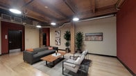 This thoughtfully designed single-floor space masterfully combines industrial character with modern sophistication. The standout architectural feature is the exposed wood-plank ceiling, accented by heavy timber beams and visible ductwork, lending the space a warm, loft-like ambiance that feels both refined and approachable.

The main lounge area is anchored by a sleek, dark gray sofa with accent pillows in rich terracotta tones, paired with a live-edge wood coffee table that adds an organic, artisanal touch. Complementing the seating arrangement are two contemporary light gray armchairs with slim black metal frames, creating an inviting conversational setting. The light hardwood flooring throughout brightens the interior and provides a clean contrast to the heavier ceiling elements above.

Structural columns with exposed brick bases punctuate the space, reinforcing its industrial heritage while contributing to its visual rhythm. Tall, lush indoor plants are strategically placed to introduce natural life and color. Curated wall art adds cultural and visual interest to the neutral-toned walls.

Track and pendant lighting provide layered illumination, ensuring the space feels well-lit and welcoming at all times. Overall, the space strikes a compelling balance between raw architectural texture and polished, contemporary design.