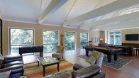 Located at 1933 North Hills Dr in Raleigh, NC, this single-story space makes a strong architectural impression with its vaulted ceiling accented by exposed white beams that draw the eye upward and create an airy, open atmosphere. The room is generously sized, offering a seamless blend of entertainment and relaxation areas beneath the soaring roofline.

Large black-framed windows line the walls, flooding the interior with natural light and framing lush, mature tree views that bring a sense of the outdoors inside. The warm hardwood flooring anchors the space and complements the neutral beige tones of the walls.

The layout is thoughtfully designed to accommodate multiple uses. A seating area features leather sofas and upholstered accent chairs arranged around a glass-top coffee table atop layered area rugs, creating an inviting lounge environment. The entertainment side of the room includes a foosball table and a full-sized billiards table, making it well-suited for social gatherings.

Overall, the space strikes a balance between casual comfort and active entertainment, enhanced by its dramatic ceiling architecture and strong connection to the surrounding natural landscape.