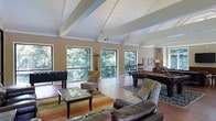 Located at 1933 North Hills Dr in Raleigh, NC, this single-story space makes a strong architectural impression with its vaulted ceiling accented by exposed white beams that draw the eye upward and create an airy, open atmosphere. The room is generously sized, offering a seamless blend of entertainment and relaxation areas beneath the soaring roofline.

Large black-framed windows line the walls, flooding the interior with natural light and framing lush, mature tree views that bring a sense of the outdoors inside. The warm hardwood flooring anchors the space and complements the neutral beige tones of the walls.

The layout is thoughtfully designed to accommodate multiple uses. A seating area features leather sofas and upholstered accent chairs arranged around a glass-top coffee table atop layered area rugs, creating an inviting lounge environment. The entertainment side of the room includes a foosball table and a full-sized billiards table, making it well-suited for social gatherings.

Overall, the space strikes a balance between casual comfort and active entertainment, enhanced by its dramatic ceiling architecture and strong connection to the surrounding natural landscape.