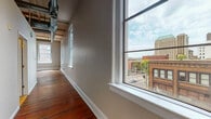 This one-bedroom, one-bathroom unit showcases an industrial-loft aesthetic rooted in historic building character. Original hardwood floors with a rich, deep patina run throughout the living spaces and bedroom, lending warmth and authenticity to the open layout. The main living and kitchen area is expansive, anchored by tall ceilings with exposed structural beams and visible ductwork that reinforce the building's heritage. A ceiling fan provides airflow across the generous open floor plan.

The kitchen is well-appointed with white cabinetry, a classic subway tile backsplash, granite countertops extending into a peninsula-style breakfast bar, and stainless steel appliances including a range and dishwasher. Large arched windows flood the living area with natural light and frame views of the surrounding urban streetscape, including neighboring historic brick buildings.

The bedroom is a comfortable, well-proportioned room also featuring hardwood floors, a ceiling fan, and additional arched windows that continue the city views. The full bathroom includes tile flooring, a tub/shower combination, and granite countertop detail at the vanity. The overall ambiance is that of a character-rich urban space blending historic architectural elements with modern finishes.