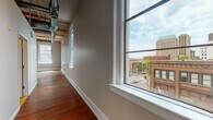 This one-bedroom, one-bathroom unit showcases an industrial-loft aesthetic rooted in historic building character. Original hardwood floors with a rich, deep patina run throughout the living spaces and bedroom, lending warmth and authenticity to the open layout. The main living and kitchen area is expansive, anchored by tall ceilings with exposed structural beams and visible ductwork that reinforce the building's heritage. A ceiling fan provides airflow across the generous open floor plan.

The kitchen is well-appointed with white cabinetry, a classic subway tile backsplash, granite countertops extending into a peninsula-style breakfast bar, and stainless steel appliances including a range and dishwasher. Large arched windows flood the living area with natural light and frame views of the surrounding urban streetscape, including neighboring historic brick buildings.

The bedroom is a comfortable, well-proportioned room also featuring hardwood floors, a ceiling fan, and additional arched windows that continue the city views. The full bathroom includes tile flooring, a tub/shower combination, and granite countertop detail at the vanity. The overall ambiance is that of a character-rich urban space blending historic architectural elements with modern finishes.