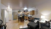 This single-floor space showcases an open-concept design that seamlessly integrates the kitchen, dining, and living areas into one cohesive environment. The kitchen is well-appointed with medium-toned wood cabinetry extending to the upper walls, offering generous storage. Stainless steel appliances, including a refrigerator, microwave, and range, provide a modern touch, while dark granite-style countertops add contrast. A small desk or workspace area is neatly positioned along the kitchen wall, adding functional versatility.

The dining area sits centrally, anchored by a dark wood table surrounded by matching chairs, illuminated by a pendant light overhead. Fresh flowers on the table contribute a welcoming detail. Light hardwood flooring flows continuously throughout, enhancing the sense of openness and warmth across the space.

The living area transitions naturally from the dining zone, featuring a charcoal upholstered sectional with decorative throw pillows, grounding the space with a comfortable, casual ambiance. A decorative wall accent adds a subtle artistic element. Natural and ambient lighting together create a bright, inviting atmosphere throughout this well-organized, multi-functional single-level space.