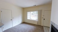 The Sandusky is a one-bedroom, one-bathroom unit designed for comfortable, efficient living on a single floor. The living area features wall-to-wall carpeting in a neutral tone, contributing to a warm and understated ambiance throughout the space. Cream-colored walls keep the interior feeling bright and open, while a sliding vertical blind panel and a large sliding glass door or wide window along one wall allow natural light to flow freely into the room, providing views of the surrounding exterior. A standard entry door is positioned nearby, offering a straightforward and accessible entryway. The ceiling features a centrally located HVAC vent, indicating a climate control system integrated throughout the unit. Tile flooring is visible at the entryway transition, providing a practical surface near the door. The overall layout is clean and uncluttered, offering a blank canvas well-suited for personalization. The unit's modest footprint is thoughtfully arranged to maximize usability, making it a practical option for individuals or couples seeking a manageable, low-maintenance living space.