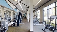 This single-floor fitness center offers a comprehensive workout environment designed to accommodate a variety of exercise needs. The space is generously sized, with an open layout that provides ample room for movement between equipment stations. Structural columns are strategically placed throughout, subtly dividing the floor into functional zones without disrupting the overall sense of openness.

The strength training area features a selection of weight machines, including a prominent cable/resistance unit and a flat bench with a barbell setup, catering to users at different fitness levels. On the cardio side, elliptical trainers, rowing machines, and additional cardiovascular equipment are arranged along the perimeter, positioned to take advantage of the large, multi-pane windows that line the exterior wall. These windows flood the space with natural light and provide views of the surrounding greenery, creating an energizing atmosphere.

Underfoot, a patterned carpet covers much of the exercise floor, while ceiling fans and rows of modern linear LED lighting ensure the space remains comfortable and well-illuminated. Full-length mirrors line one wall, enhancing the sense of depth and allowing users to monitor their form. The overall aesthetic is clean, functional, and modern, making this fitness center a well-rounded facility for daily training.