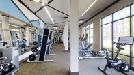 This single-floor fitness center offers a comprehensive workout environment designed to accommodate a variety of exercise needs. The space is generously sized, with an open layout that provides ample room for movement between equipment stations. Structural columns are strategically placed throughout, subtly dividing the floor into functional zones without disrupting the overall sense of openness.

The strength training area features a selection of weight machines, including a prominent cable/resistance unit and a flat bench with a barbell setup, catering to users at different fitness levels. On the cardio side, elliptical trainers, rowing machines, and additional cardiovascular equipment are arranged along the perimeter, positioned to take advantage of the large, multi-pane windows that line the exterior wall. These windows flood the space with natural light and provide views of the surrounding greenery, creating an energizing atmosphere.

Underfoot, a patterned carpet covers much of the exercise floor, while ceiling fans and rows of modern linear LED lighting ensure the space remains comfortable and well-illuminated. Full-length mirrors line one wall, enhancing the sense of depth and allowing users to monitor their form. The overall aesthetic is clean, functional, and modern, making this fitness center a well-rounded facility for daily training.