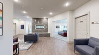 Located at Heritage Towne Blvd in Midlothian, Texas, this contemporary community lounge offers a generously sized, single-floor gathering space designed for comfort and social interaction. The open-concept layout is anchored by a striking floor-to-ceiling textured brick fireplace feature wall with a sleek linear electric fireplace insert, serving as a focal point that elegantly divides the space into distinct yet connected seating areas. Throughout the lounge, warm wood-look flooring complements the crisp white walls, creating a clean and welcoming aesthetic. Multiple seating arrangements—featuring deep blue sofas and rich cognac leather chairs—are thoughtfully positioned atop patterned area rugs, accommodating both larger gatherings and more intimate conversations. Recessed lighting and modern pendant and multi-arm fixtures illuminate the space, while curated wall art adds pops of color and personality. A dedicated kitchenette area is equipped with gray cabinetry, a sink, open wood shelving, and a coffee station, with a large window framing views of the outdoors. French doors lead to a private balcony enclosed by contemporary horizontal-rail black metal railings, offering serene views of the surrounding natural landscape. The overall ambiance is modern, polished, and inviting—ideal for community gatherings.