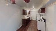 This single-floor unit offers a straightforward, functional layout with a clear separation between living and sleeping areas. The kitchen is equipped with dark wood cabinetry, white appliances including a coil-burner range and refrigerator, a double-basin sink, and light countertops. Durable hardwood-style flooring runs through the kitchen and adjoining living area, while a ceiling fan overhead provides added ventilation.

The bedrooms are carpeted in a neutral gray tone and feature built-in closets, including one room with an open shelving niche and a built-in desk surface, making it well-suited for use as a home office or study. Each bedroom is finished with simple flush-mount lighting and white walls that contribute to a clean, neutral aesthetic. Natural light enters through slatted blinds on the windows.

The bathroom includes a vanity with a light countertop, a towel bar, and a tub-shower combination, all finished in white. The 3D floor plan illustrates a compact but efficient layout connecting the kitchen, living space, and bedrooms in a logical flow. Overall, the unit maintains a clean, move-in-ready appearance with consistent neutral finishes throughout.