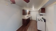 This single-floor unit offers a straightforward, functional layout with a clear separation between living and sleeping areas. The kitchen is equipped with dark wood cabinetry, white appliances including a coil-burner range and refrigerator, a double-basin sink, and light countertops. Durable hardwood-style flooring runs through the kitchen and adjoining living area, while a ceiling fan overhead provides added ventilation.

The bedrooms are carpeted in a neutral gray tone and feature built-in closets, including one room with an open shelving niche and a built-in desk surface, making it well-suited for use as a home office or study. Each bedroom is finished with simple flush-mount lighting and white walls that contribute to a clean, neutral aesthetic. Natural light enters through slatted blinds on the windows.

The bathroom includes a vanity with a light countertop, a towel bar, and a tub-shower combination, all finished in white. The 3D floor plan illustrates a compact but efficient layout connecting the kitchen, living space, and bedrooms in a logical flow. Overall, the unit maintains a clean, move-in-ready appearance with consistent neutral finishes throughout.