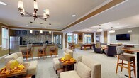 This single-floor community clubhouse is thoughtfully designed to foster social interaction and leisure. The main lounge area is an open, generously proportioned space anchored by plush patterned armchairs and sofas arranged around a central coffee table, all bathed in warm light from a striking multi-arm chandelier and pendant fixtures. Warm taupe and gold tones on the walls, complemented by white crown molding and decorative columns, lend the space an inviting, upscale ambiance.

A wet bar and refreshment counter runs along one wall, featuring dark granite countertops, warm wood cabinetry, a mosaic tile backsplash, and bar-height seating — ideal for casual gatherings. The adjacent full kitchen mirrors this aesthetic with matching cabinetry, granite surfaces, stainless steel appliances, and tile flooring, providing ample prep space for events.

The game room area includes a pool table and a classic shuffleboard table, complemented by a wall-mounted flat-screen TV and comfortable seating. High-top bistro tables throughout offer additional casual dining options.

A welcoming tiled entryway with a decorative console table and prominent signage directs guests to the on-site fitness center. Throughout the clubhouse, large windows invite natural light, creating a bright and comfortable environment suited for community gatherings year-round.