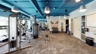 This contemporary fitness center is designed to deliver a comprehensive workout experience across a single, open floor. The space is immediately distinguished by its striking teal-painted exposed ceiling, complete with visible ductwork and pendant lighting that lends an industrial-chic aesthetic. Large floor-to-ceiling windows line one side of the cardio zone, flooding the area with natural light and offering views of an outdoor patio with seating and umbrellas, creating an energizing atmosphere for workouts.

The cardio section is outfitted with a full row of Life Fitness elliptical trainers and treadmills, each equipped with individual display screens. The strength training area features a well-curated selection of cable machines, weight benches, and a tiered dumbbell rack positioned beneath a bold large-scale portrait mural that serves as a dramatic focal point. Full-length mirrors extend depth throughout the space.

A dedicated studio room, accessible through sliding textured panel doors, provides space for functional training with TRX suspension straps, a spin bike, and open floor area ideal for group classes or individual sessions. The flooring throughout transitions between patterned carpet tiles and hard surfaces, offering both comfort and functionality. Overall, the space combines vibrant design elements with a well-rounded equipment offering suited for diverse fitness needs.