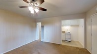 This single-floor residence offers a straightforward and functional layout designed for comfortable everyday living. The main living area is finished with wall-to-wall carpeting in a warm neutral tone, creating a cozy atmosphere throughout. A ceiling fan with an integrated light fixture is centrally mounted, providing both air circulation and ambient lighting to the space.

The living area flows openly toward the kitchen, which is tucked into an alcove and outfitted with white upper cabinetry and matching white appliances, including a freestanding gas range. The kitchen is finished with a light-toned floor, offering a clean contrast to the carpeted living space. Under-cabinet lighting adds a practical brightness to the cooking area.

A doorway off the main living area leads to what appears to be a tiled bathroom or utility space, partially visible and featuring patterned tile work. The walls throughout are painted in a soft, neutral tone, contributing to the overall bright and airy feel of the interior.

The space is clean, well-maintained, and efficiently arranged, making it well-suited for those seeking a simple, low-maintenance living environment. The open sightlines between the living and kitchen areas enhance the sense of space and connectivity throughout the single-floor layout.